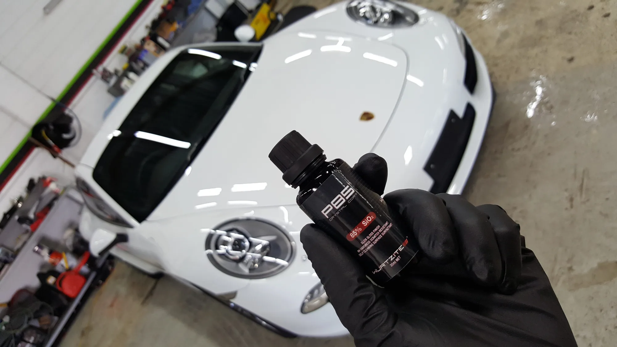 Gloved hand holding a bottle of Wurtzite P85 beside a white Porsche 911